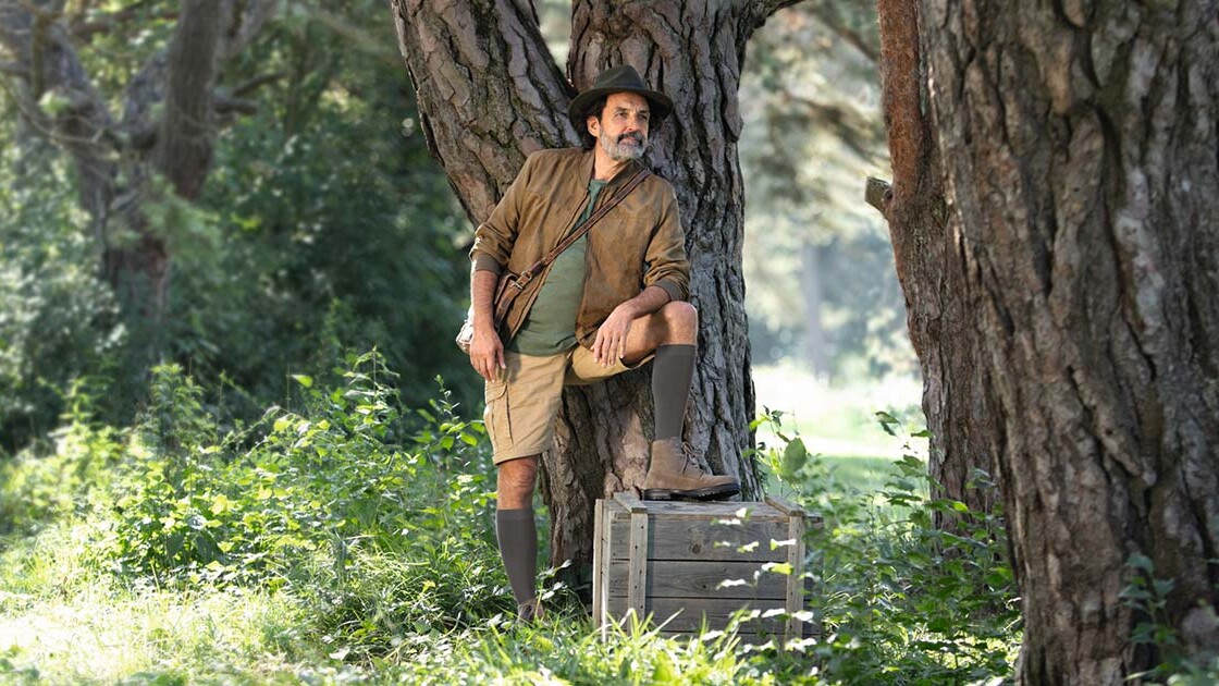 Ein Mann mit Entdecker-Outfit steht in der Natur und schaut in die Ferne. Er trägt kniehohe Kompressionsstrümpfe. Ein Mann mit Entdecker-Outfit steht in der Natur und schaut in die Ferne. Er trägt kniehohe Kompressionsstrümpfe.