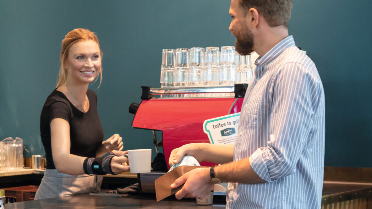 A waitress wearing a JuzoFlex Manu Xtra wrist support hands a cup to a customer over the counter