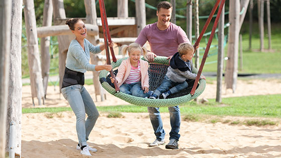 A family at the playground. The children are on the swings. The mother is wearing a back brace