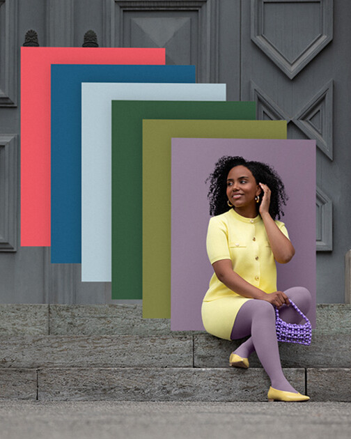 Una mujer con medias de compresión en color Cozy Violet está sentada en unas escaleras. Al fondo, los paneles de color de la actual colección Juzo Trend Colours.